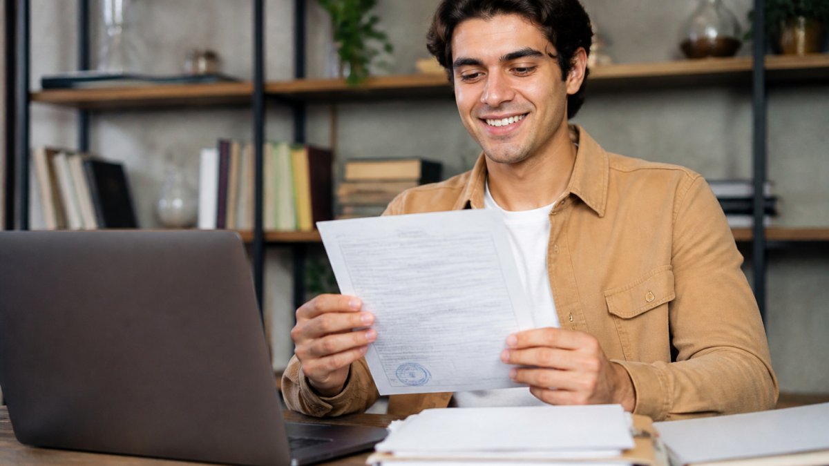 Joven profesional revisando documentos oficiales en un escritorio con computadora portátil, sosteniendo un acta de nacimiento y sonriendo con confianza